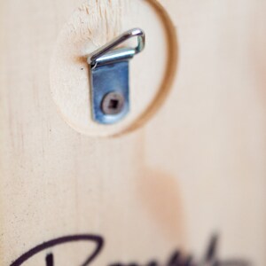 May include: A close-up of a wooden surface with a metal hanging hook attached. The hook is silver and has a small screw in the center. The wood is light brown and has a natural grain.
