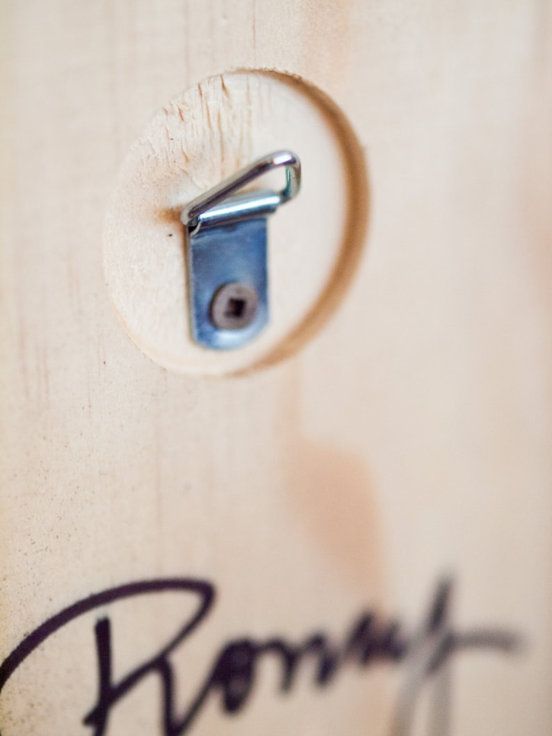 May include: A close-up of a wooden surface with a metal hanging hook attached. The hook is silver and has a small screw in the center. The wood is light brown and has a natural grain.