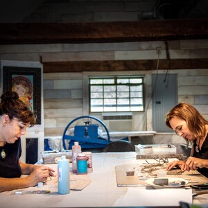 May include: Two people are working on mosaic art projects at a table. They are using small tiles and tools to create their designs. There are bottles of paint and other supplies on the table.