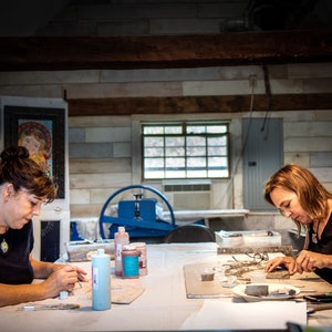 May include: Two people are working on mosaic art projects at a table. They are using small pieces of tile and glass to create their designs. There are bottles of glue and other supplies on the table.