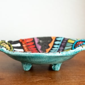 May include: A teal ceramic bowl with a colorful geometric design and three legs. The bowl is decorated with black, white, red, orange, yellow, green, blue, and purple.