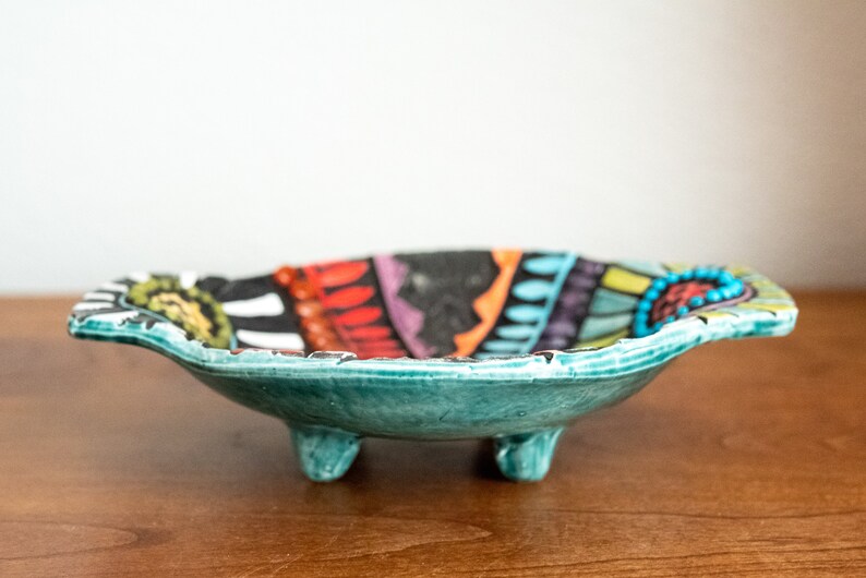 May include: A teal ceramic bowl with a colorful geometric design and three legs. The bowl is decorated with black, white, red, orange, yellow, green, blue, and purple.