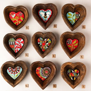 May include: Nine wooden heart-shaped bowls with different colorful painted designs. The designs include a bee, a flower, a mosaic, and a geometric pattern. The bowls are arranged in three rows of three.