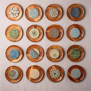 May include: A collection of sixteen circular wooden coasters, each featuring a unique ceramic inlay with distinct geometric, floral, or abstract patterns in various colors including blue, green, gray, and beige, arranged in a four-by-four grid.
