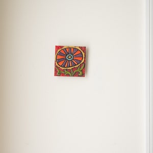 May include: A colorful ceramic tile with a stylized flower design. The flower is red, orange, purple, and blue with a green stem and leaves.