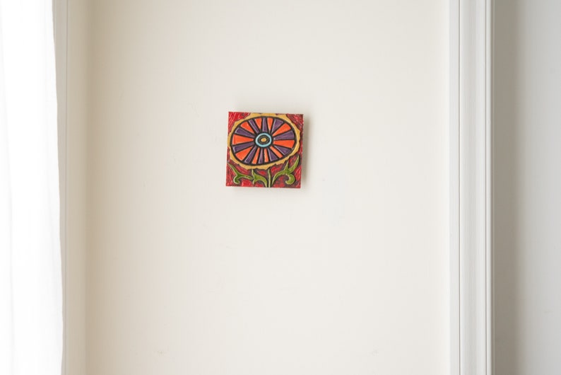 May include: A colorful ceramic tile with a stylized flower design. The flower is red, orange, purple, and blue with a green stem and leaves.