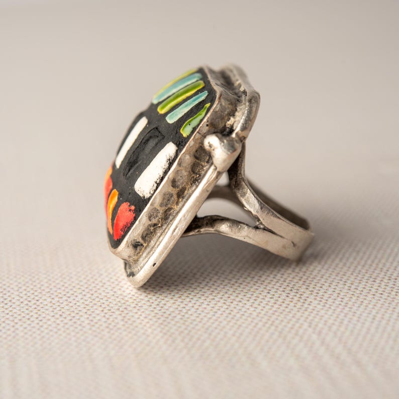 A silver ring with a square shaped ceramic tile top. The tile is decorated with a colorful tile in shades of red, orange, green, blue, black and white. Handmade in USA by ceramic and mosaic artists Romy and Clare, Jasper, IN.