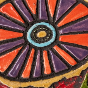May include: A close-up of a colorful ceramic tile with a circular design. The center of the tile features a blue, turquoise, and yellow circle with a black outline. The circle is surrounded by alternating orange and purple stripes.