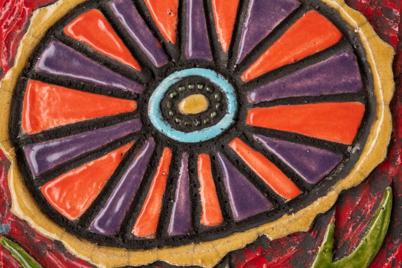 May include: A close-up of a colorful ceramic tile with a circular design. The center of the tile features a blue, turquoise, and yellow circle with a black outline. The circle is surrounded by alternating orange and purple stripes.