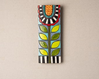 Könnte beinhalten: Ein farbenfroher Wandbehang aus Keramik mit einem stilisierten Blumendesign. Die Blume hat eine rote Mitte, gelbe Blütenblätter und einen blau-weiß gestreiften Stiel. Die Blätter sind grün und gelb.