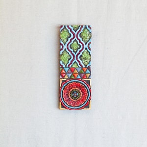 May include: A colorful tile with a geometric pattern in shades of green, blue, red, and yellow. The tile has a circular design in the center with a cross in the middle.