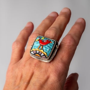 May include: A square silver ring featuring a mosaic inlay of a red bird against a textured light blue background, with a yellow sun and black and white geometric accents at the bottom, set in a polished silver frame with a wide, sturdy band.
