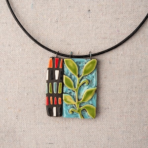 Puede incluir: Un collar de cordón negro con un colgante de cerámica que presenta un patrón geométrico en negro, blanco, rojo y verde en un lado y una rama verde con hojas sobre un fondo azul en el otro.