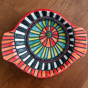 May include: A red ceramic dish with a colorful mosaic design. The center of the dish features a circular pattern of black, white, green, blue, and orange stripes. The outer edge of the dish is decorated with a band of red, orange, and black stripes.