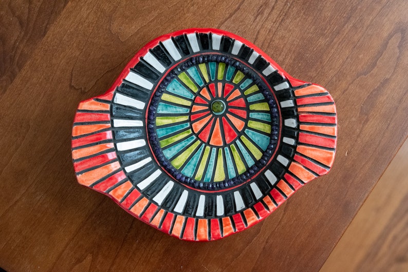 May include: A red ceramic dish with a colorful mosaic design. The center of the dish features a circular pattern of black, white, green, blue, and orange stripes. The outer edge of the dish is decorated with a band of red, orange, and black stripes.
