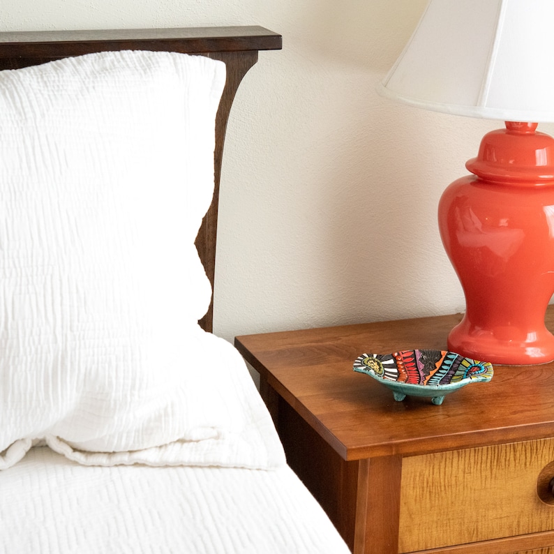 May include: A white bed with a white pillow and a wooden headboard. A red lamp with a white shade is on a wooden nightstand. A small, colorful ceramic dish with three legs is on the nightstand.