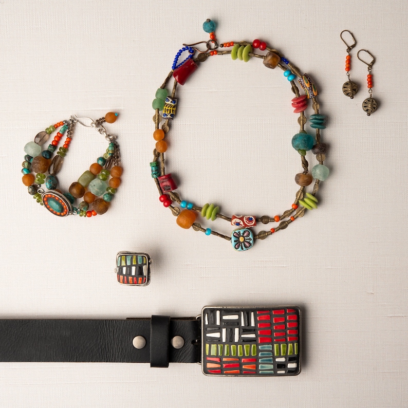 May include: A set of colorful jewelry featuring a necklace, bracelet, earrings, and a belt buckle. The necklace and bracelet are made of various beads in shades of green, blue, red, and orange. The earrings are simple silver hoops with small beads hanging from them. The belt buckle is a square piece of metal with a mosaic design in shades of red, green, blue, and white.
