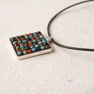 May include: A square pendant necklace with a silver frame and a mosaic of colorful stones. The necklace is on a black leather cord.