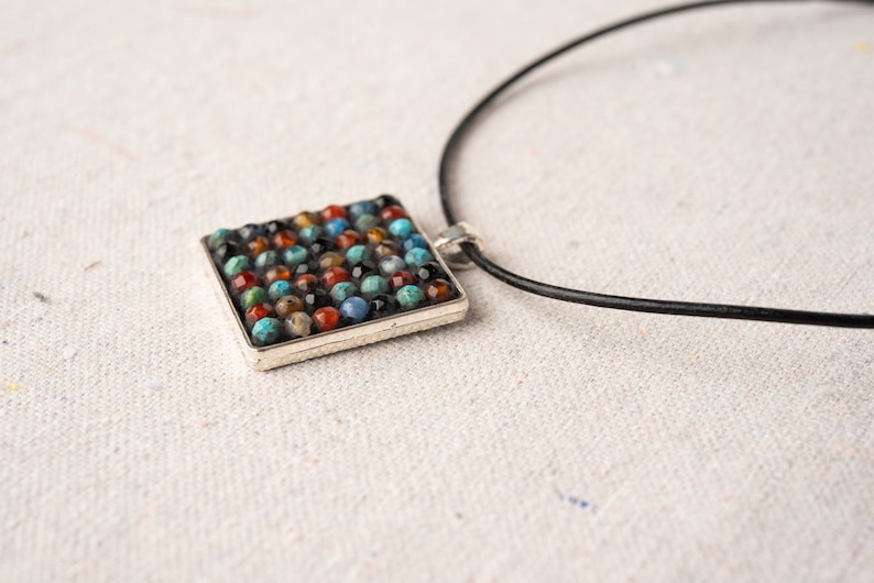 May include: A square pendant necklace with a silver frame and a mosaic of colorful stones. The necklace is on a black leather cord.