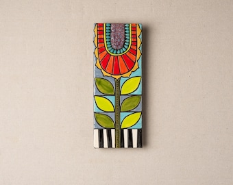 Könnte beinhalten: Eine farbenfrohe Keramikfliese mit einem stilisierten Blumendesign. Die Blume ist rot, orange und gelb mit einem grünen Stiel und Blättern. Die Fliese hat einen schwarz-weißen Streifenrand.
