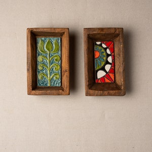 May include: Two wooden rectangular trays with decorative ceramic tiles. The tile on the left tray features a green and blue stylised plant design. The tile on the right tray features a colourful geometric design with red, orange, yellow, blue, green, and white.