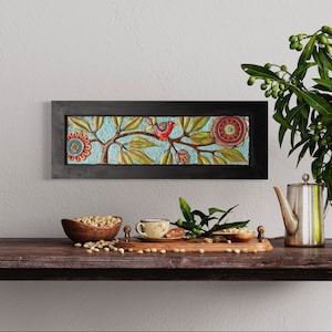 Puede incluir: Una obra de arte enmarcada con un fondo azul que presenta un árbol estilizado con flores rojas y un pájaro. La obra de arte está en un marco negro y está colgada en una pared blanca sobre una mesa de madera.