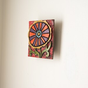 May include: A colorful ceramic wall art piece featuring a stylized flower with orange, purple, and yellow petals. The flower has a green stem and leaves. The background is a red and brown textured surface.