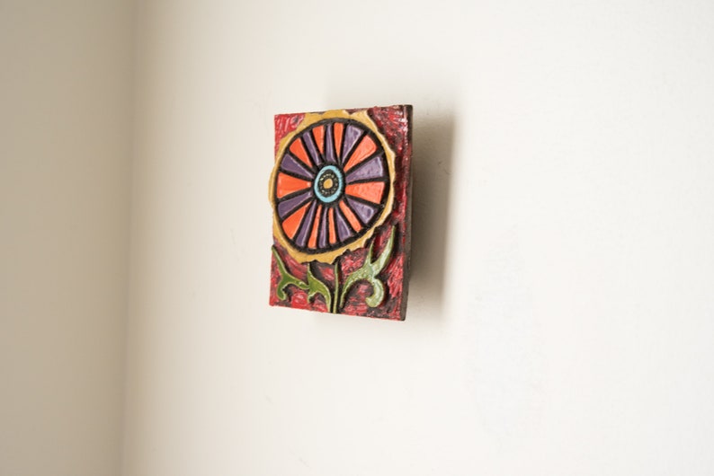 May include: A colorful ceramic wall art piece featuring a stylized flower with orange, purple, and yellow petals. The flower has a green stem and leaves. The background is a red and brown textured surface.