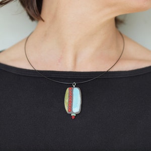 May include: A black cord necklace with a rectangular pendant. The pendant is made of gray, blue, green, and red clay with a geometric design.