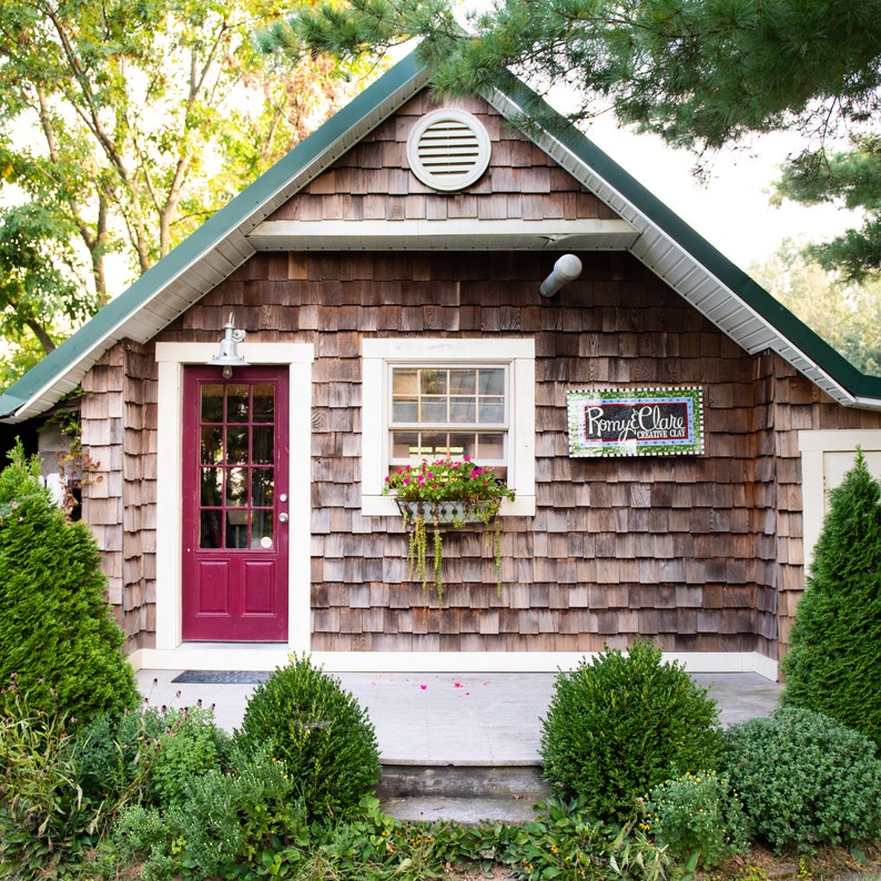 May include: A small, wooden building with a red door and a window with a flower box. The building has a sign that says "Rommy & Claire Creative Clay".