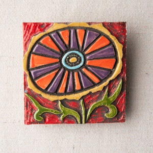 May include: A ceramic tile with a colorful flower design. The flower is orange, purple, and yellow with a blue and black center. The tile is set against a red background.
