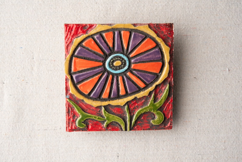 May include: A ceramic tile with a colorful flower design. The flower is orange, purple, and yellow with a blue and black center. The tile is set against a red background.