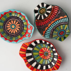 May include: Three ceramic dishes with colorful mosaic designs. The dishes are oval shaped and have a raised rim. The designs feature geometric patterns in shades of red, orange, yellow, green, blue, black, and white.