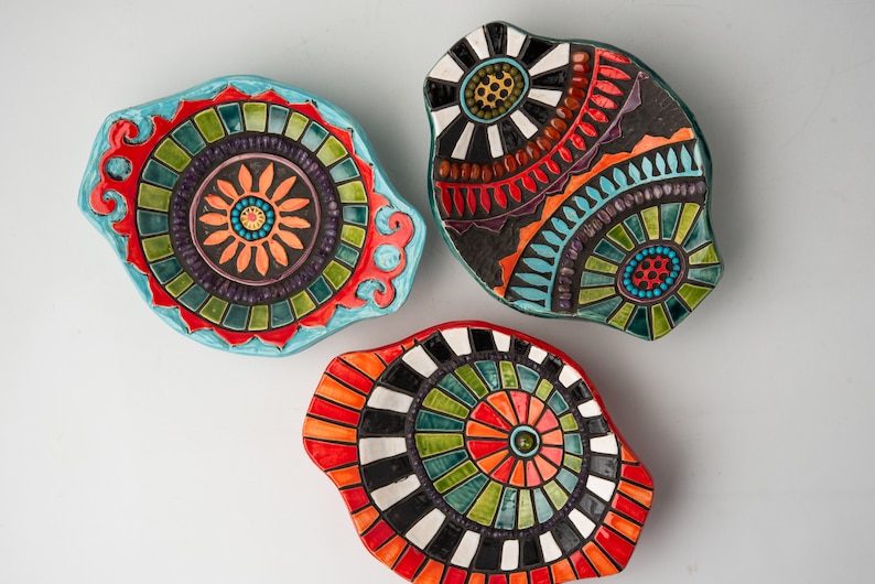 May include: Three ceramic dishes with colorful mosaic designs. The dishes are oval shaped and have a raised rim. The designs feature geometric patterns in shades of red, orange, yellow, green, blue, black, and white.