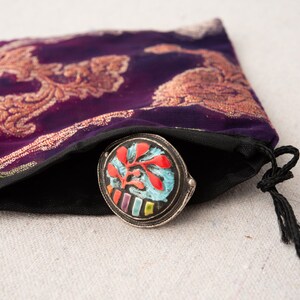Pode incluir: Uma pequena caixa redonda de prata com um design floral colorido. A caixa est&aacute; dentro de uma bolsa de veludo roxo com cord&atilde;o e bordados dourados.