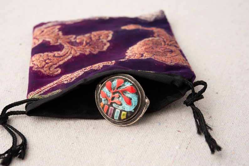Pode incluir: Uma pequena caixa redonda de prata com um design floral colorido. A caixa est&aacute; dentro de uma bolsa de veludo roxo com cord&atilde;o e bordados dourados.