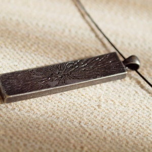 May include: A silver pendant with a textured, rectangular design. The pendant is attached to a black cord.