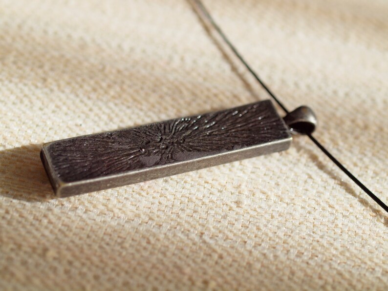May include: A silver pendant with a textured, rectangular design. The pendant is attached to a black cord.