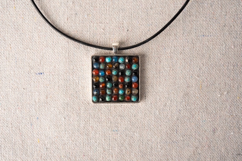 May include: A square pendant necklace with a black leather cord. The pendant is made of silver and features a mosaic of colorful gemstones.