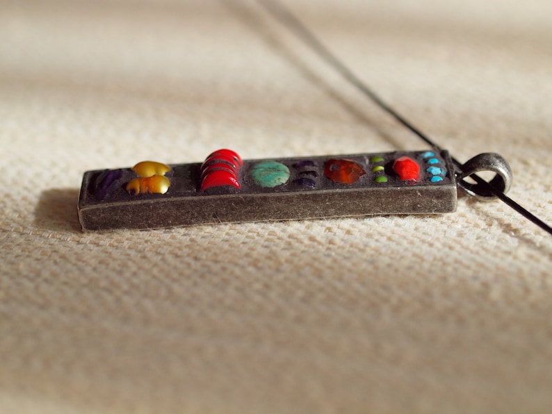 May include: A silver pendant with a rectangular shape and a variety of colorful stones. The stones are arranged in a row and include red, yellow, green, blue, and purple.
