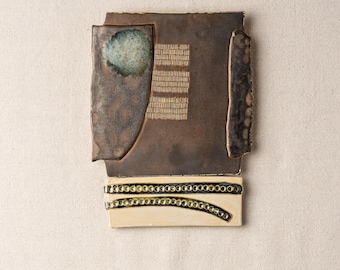 May include: Abstract ceramic wall art with a layered design. The piece features a dark brown rectangular base with a teal-colored accent and textured details. A cream-colored section at the bottom has a beaded design.