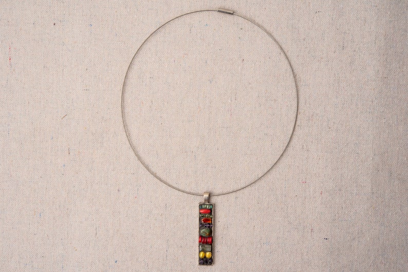 Puede incluir: Un collar de plata con un colgante hecho de azulejos de mosaico de colores. El colgante es rectangular y presenta azulejos rojos, verdes, amarillos y negros.
