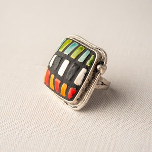 A silver ring with a square shaped ceramic tile top. The tile is decorated with a colorful tile in shades of red, orange, green, blue, black and white. Handmade in USA by ceramic and mosaic artists Romy and Clare, Jasper, IN.