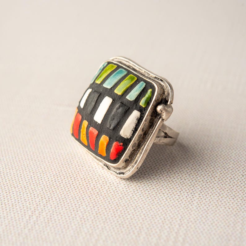 A silver ring with a square shaped ceramic tile top. The tile is decorated with a colorful tile in shades of red, orange, green, blue, black and white. Handmade in USA by ceramic and mosaic artists Romy and Clare, Jasper, IN.