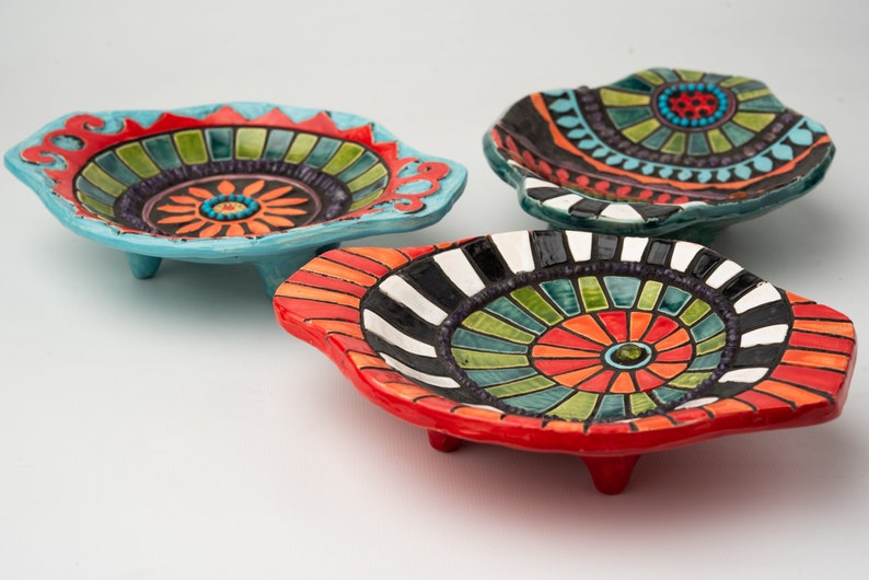 May include: Three colorful ceramic bowls with geometric patterns and raised edges. The bowls are decorated with bright colors, including red, orange, yellow, green, blue, and black. Each bowl has three small feet.