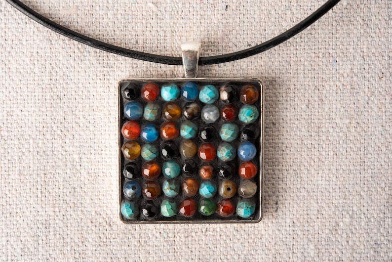 May include: A square pendant necklace with a silver frame and a mosaic of colorful stones. The stones are set in a grid pattern and include shades of blue, green, red, and brown.