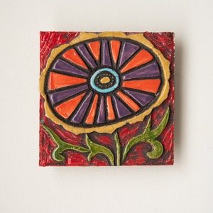 May include: A ceramic tile with a colorful flower design. The flower is orange, purple, and blue with a yellow outline. The background is red.