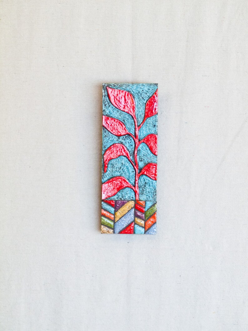 May include: A rectangular wall hanging with a blue background and a red stylized plant design. The bottom of the hanging has a colorful geometric pattern.