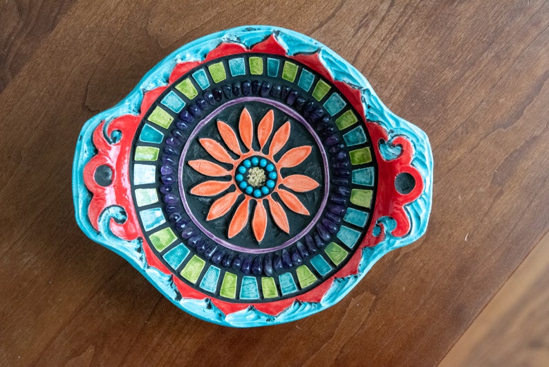 May include: A blue and red ceramic bowl with a mosaic design featuring a colorful flower in the center. The bowl has two handles on the sides.