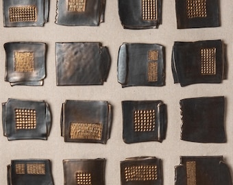 May include: A collection of sixteen dark gray, square-shaped ceramic tiles with gold-colored textured patterns. The patterns vary, including grids, lines, and irregular shapes, creating a visually interesting display. The tiles are arranged in a grid.